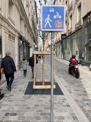 A street sign in Lyon, France shows a large image of a pedestrian, a smaller image of a cyclist, and an even smaller image of a car. 