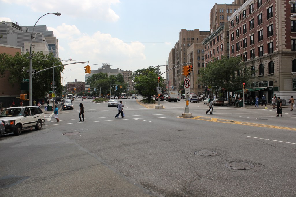 Pedestrians cross a wide intersection of streets on upper Broadway in New York City.