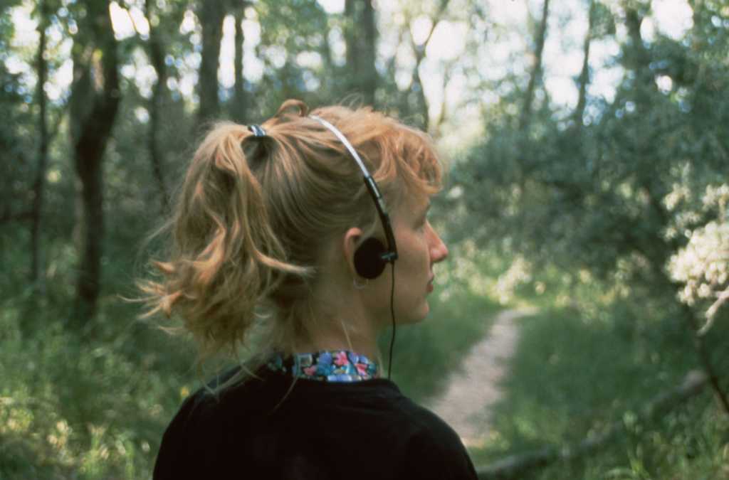 The artist Janet Cardiff walks on a path through a wood, with earphones on.